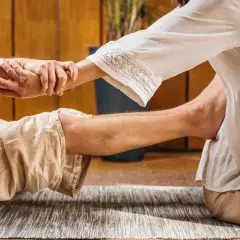 Un praticante esegue un massaggio thailandese, allungando le gambe e le braccia di un ricevente sdraiato su un tappeto.
