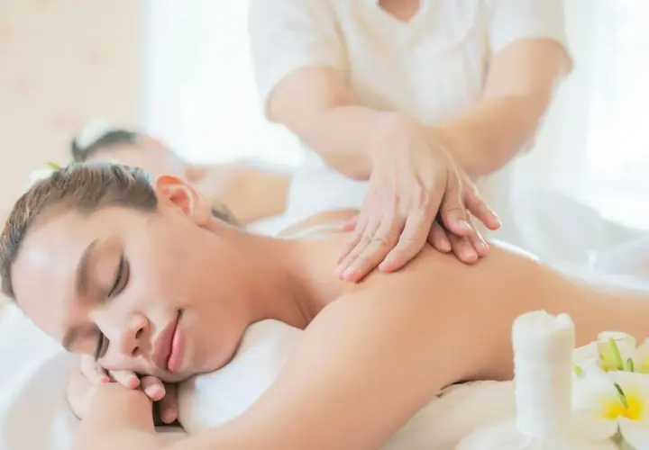 Donna rilassata riceve massaggi olistici da un terapista. Fiori bianchi e un sacchetto di erbe completano la scena di benessere.