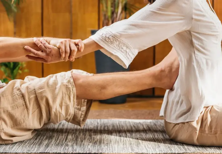 Un praticante esegue un massaggio thailandese, allungando le gambe e le braccia di un ricevente sdraiato su un tappeto.