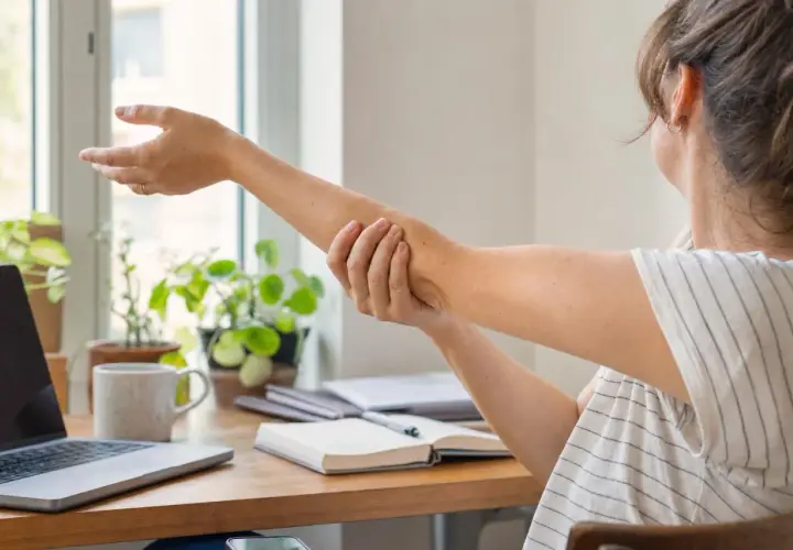 Donna si massaggia il braccio, forse per alleviare la sensazione di ritenzione idrica braccia. Sul tavolo, un laptop e piante verdi.
