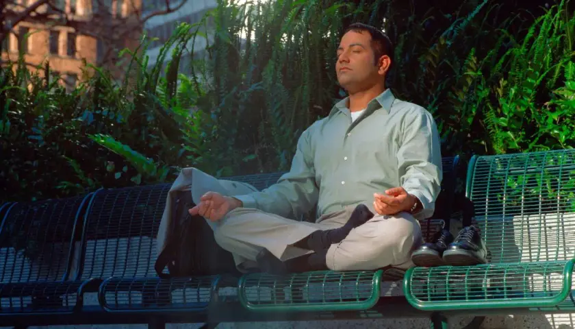 Uomo in meditazione su una panchina, con un atteggiamento zen. Le mani in posizione di preghiera, gli occhi chiusi, circondato dal verde.