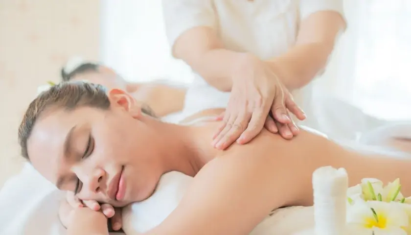 Donna rilassata riceve massaggi olistici da un terapista. Fiori bianchi e un sacchetto di erbe completano la scena di benessere.