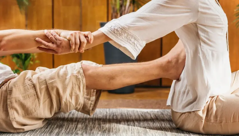 Un praticante esegue un massaggio thailandese, allungando le gambe e le braccia di un ricevente sdraiato su un tappeto.