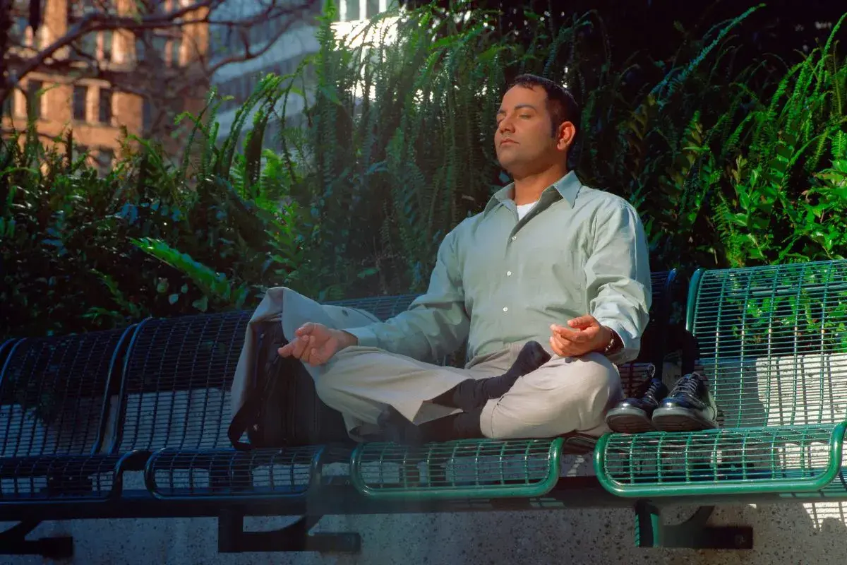Uomo in meditazione su una panchina, con un atteggiamento zen. Le mani in posizione di preghiera, gli occhi chiusi, circondato dal verde.