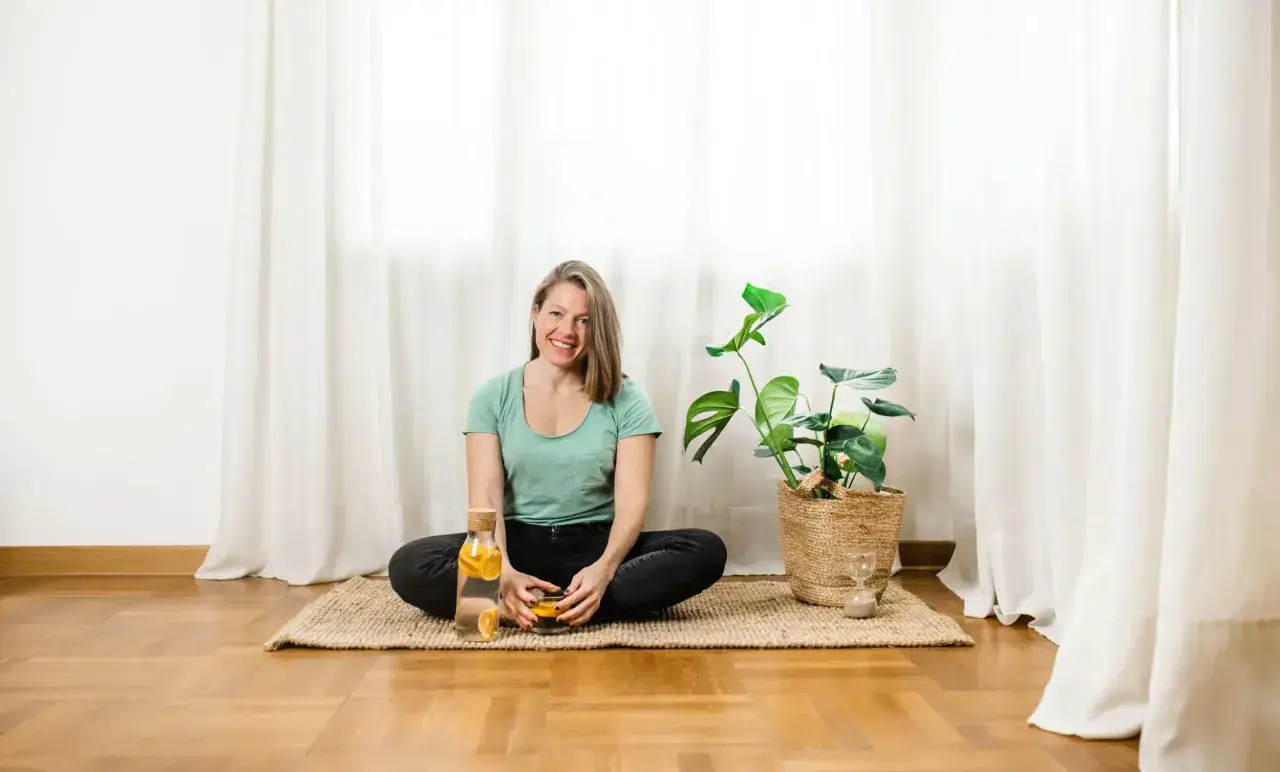 Donna sorridente in posizione yoga, con acqua aromatizzata e una pianta. Un momento di relax, sperando che il suo rene bloccato si risolva presto.