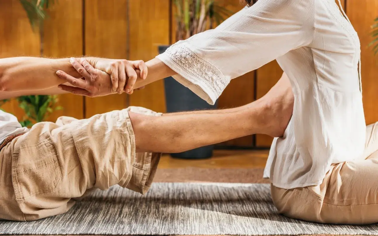 Un praticante esegue un massaggio thailandese, allungando le gambe e le braccia di un ricevente sdraiato su un tappeto.