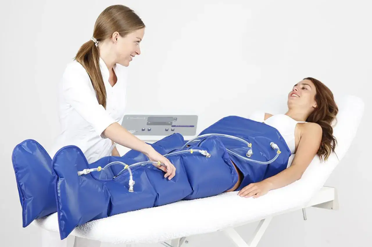 Donna sdraiata su lettino, con gambe avvolte in una tuta blu per la pressoterapia. Un'operatrice controlla il macchinario.